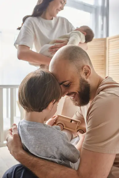 Plan vertical concentré de père jouant avec son fils avec sa femme floue avec le nouveau-né sur toile de fond — Photo de stock