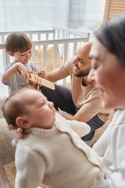 Tiro enfocado vertical del padre feliz jugando con su pequeño hijo al lado de su esposa borrosa y su bebé - foto de stock