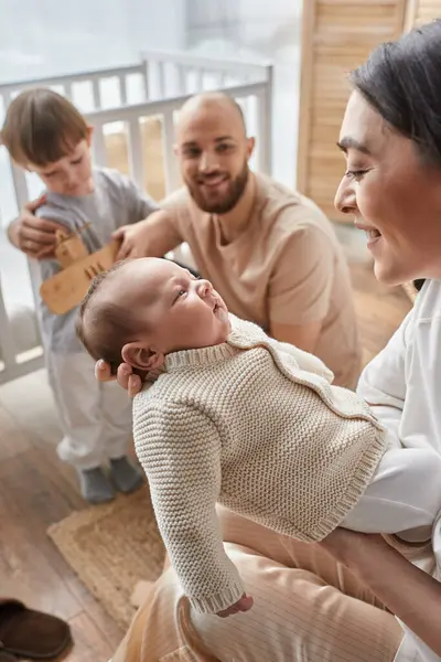 Vertical focused shot of jolly mother holding baby boy in front of blurred husband and little son — Stock Photo