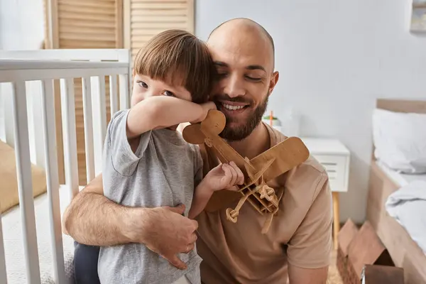 Feliz padre guapo abrazando calurosamente a su pequeño hijo mientras juega con avión de madera, concepto de familia - foto de stock