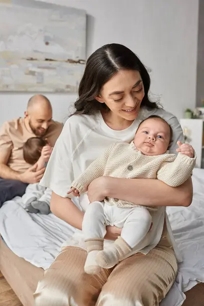 Tiro vertical de la madre sosteniendo a su bebé recién nacido con su marido borroso y su hijo pequeño en el telón de fondo - foto de stock