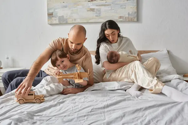 Padre barbudo jugando con su pequeño hijo al lado de su esposa sosteniendo bebé recién nacido, concepto de familia - foto de stock