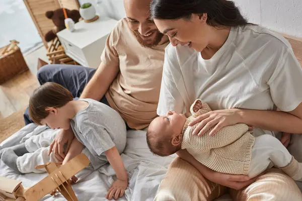 Genitori felici guardando amorevolmente il loro bambino appena nato accanto al loro piccolo figlio che gioca con i giocattoli — Foto stock
