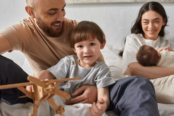 Joyful preschool boy sitting on bed surrounded by his loving parents and his newborn brother — Stock Photo