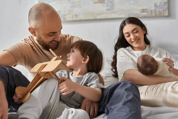 Handsome father smiling at his little son sitting on bed next to his wife holding their newborn baby — Stock Photo
