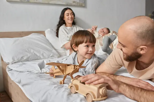 Enfoque en el padre y el pequeño hijo acostado en la cama con la madre borrosa con el bebé recién nacido en el telón de fondo - foto de stock