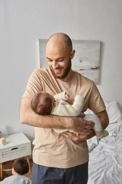 Homme barbu en tenue de maison tenant son nouveau-né et lui souriant joyeusement, concept de famille — Photo de stock
