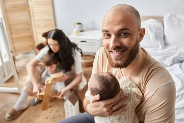 Father holding baby boy smiling at camera next to blurred wife with their little son on backdrop — Stock Photo
