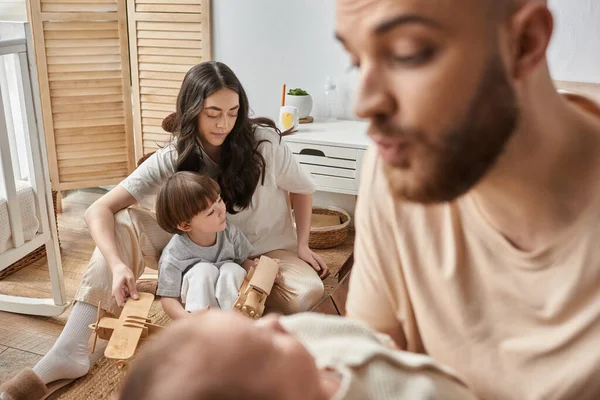 Focus on young mother sitting on floor with little son in front of blurred husband and newborn baby — Stock Photo