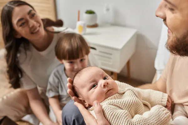Se centran en el bebé recién nacido lindo mirando a la cámara rodeada por sus padres borrosos y hermano, familia - foto de stock