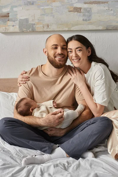 Vertikale Aufnahme freudiger Eltern, die lächelnd auf dem Bett sitzen und ihr niedliches Neugeborenes halten, Familie — Stockfoto