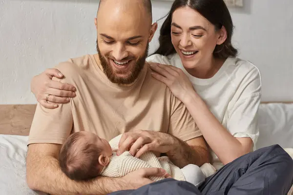 Padres modernos de buen aspecto abrazando y sosteniendo a su bebé recién nacido en brazos sonriendo alegremente, familia - foto de stock