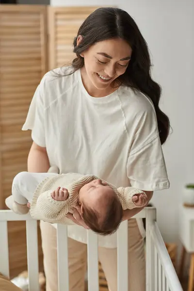 Tiro vertical de feliz madre moderna poniendo a su lindo bebé recién nacido en su cuna, concepto de familia - foto de stock