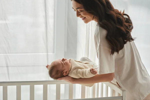 Joyous beautiful mother putting her newborn baby boy into his crib and smiling at him, family — Stock Photo
