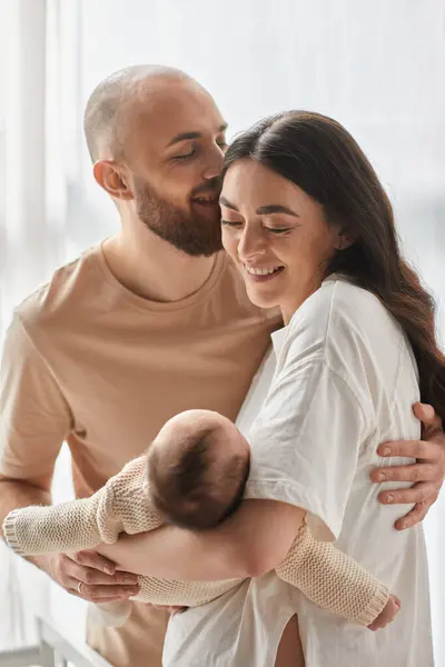 Tiro vertical de alegres padres felices abrazándose amorosamente y sosteniendo a su bebé recién nacido, familia - foto de stock