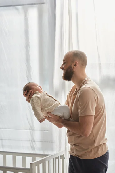 Vertical shot of happy father in cozy homewear holding his newborn son and smiling at him lovingly — Stock Photo