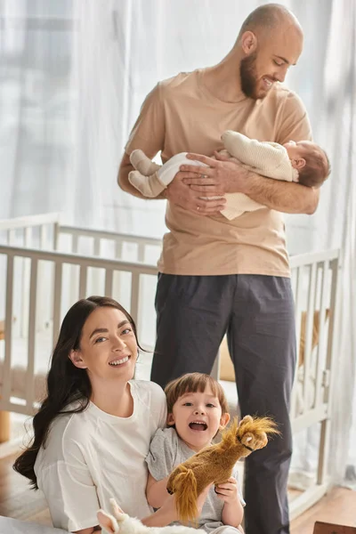 Concentre-se em mãe brincando com seu filho e sorrindo para a câmera ao lado de seu marido borrado segurando bebê — Fotografia de Stock