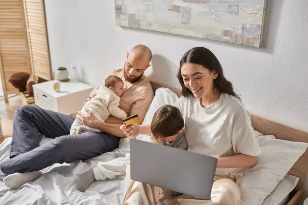 Joyous relaxing family spending time together in bed with laptop and credit card, modern parenting — Stock Photo