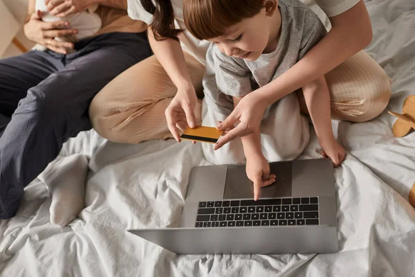 Blick von oben auf kleinen Jungen, der auf Laptop zeigt, umgeben von Familie mit seiner Mutter, die Kreditkarte hält — Stockfoto