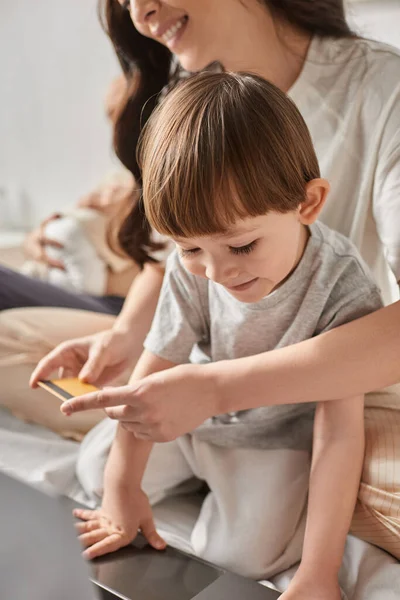 Plano enfocado vertical del niño mirando el ordenador portátil al lado de su borrosa madre sosteniendo la tarjeta de crédito - foto de stock