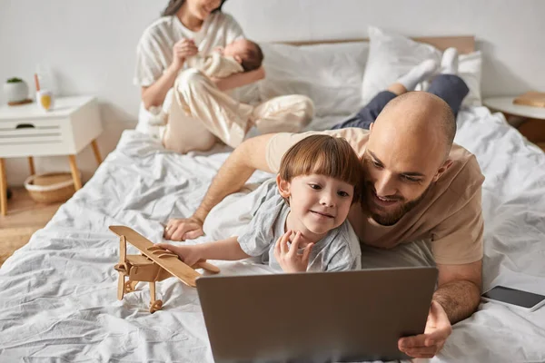 Centrarse en el niño pequeño con su padre mirando a la computadora portátil junto a su madre borrosa y su hermano pequeño - foto de stock