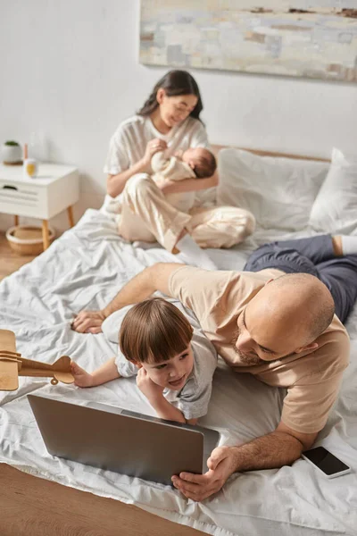 Plan vertical du père et du petit fils regardant l'ordinateur portable à côté de la mère floue tenant bébé garçon — Photo de stock