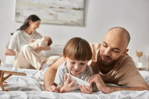 Modern father looking at phone with his little son with his blurred wife and baby on backdrop — Stock Photo