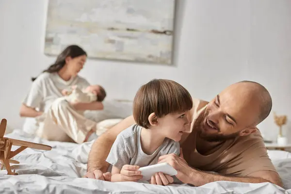 Alegre padre cariñoso sonriendo a su pequeño hijo mientras está acostado en la cama junto a su borrosa esposa y su bebé - foto de stock
