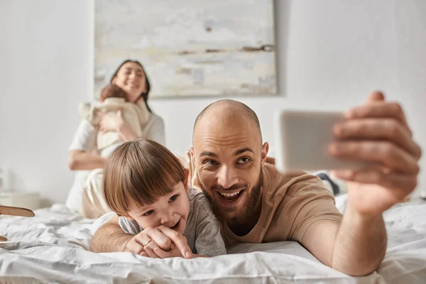 Der fröhliche Vater macht ein Selfie mit seinem Sohn und seiner verschwommenen Frau und dem Neugeborenen — Stockfoto