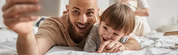 Joyous bearded father taking selfies with his little son while lying on bed, family concept, banner — Stock Photo