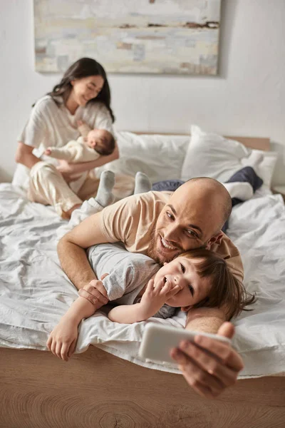 Tiro vertical de padre cariñoso tomando selfies con su pequeño hijo al lado de su borrosa esposa y bebé - foto de stock