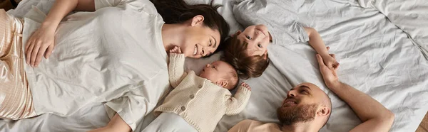 Вид сверху современной семьи в домашней одежде, лежащей на кровати и счастливо улыбающейся, современный парик, баннер — стоковое фото