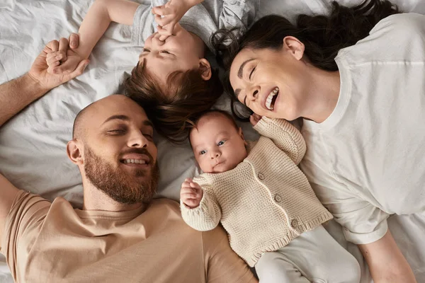 Вид сверху веселой молодой современной семьи в уютной домашней одежде, лежащей вместе на кровати и счастливо улыбающейся — стоковое фото