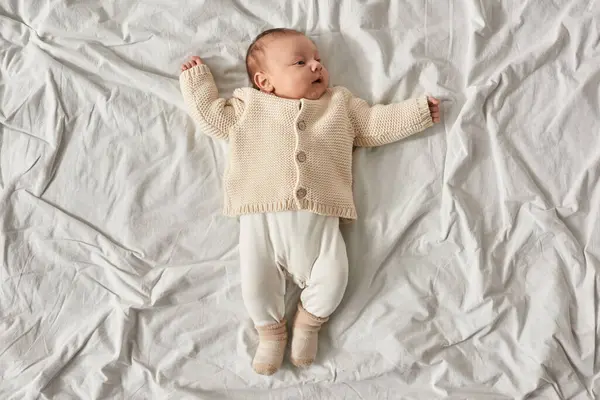 Von oben: entzückender neugeborener Junge in beige warmer Strickjacke auf weißer Decke liegend mit erhobenen Armen — Stockfoto