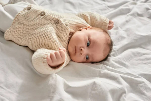 Retrato de un adorable bebé recién nacido acostado sobre una manta blanca en un cálido cárdigan beige mirando hacia otro lado - foto de stock