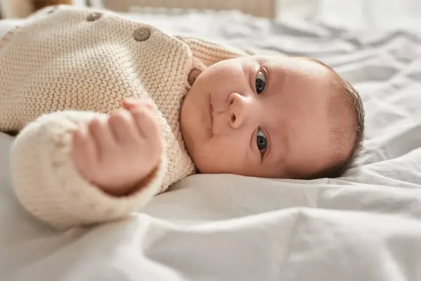 Porträt eines entzückenden kleinen neugeborenen Jungen in beiger Strickjacke, der auf dem Bett liegt und in die Kamera blickt — Stockfoto