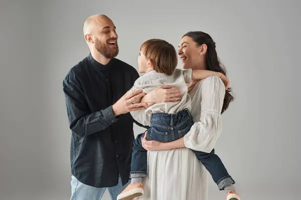Padres modernos felices en trajes elegantes sosteniendo a su pequeño hijo en el telón de fondo gris, concepto de familia - foto de stock