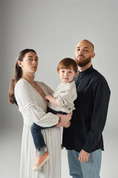 Plano vertical de los padres con estilo moderno posando con su hijo en las manos y mirando a la cámara, familia - foto de stock