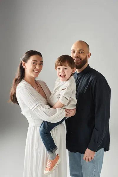 Tiro vertical de feliz familia alegre con alegre niño en las manos sobre fondo gris, familia - foto de stock