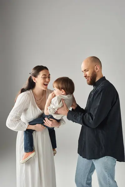 Fröhliche moderne Eltern lächeln und halten ihr kleines Kind vor grauem Hintergrund, Familienkonzept — Stockfoto