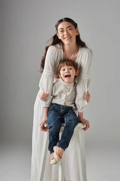 Tiro vertical de madre feliz en vestido blanco posando con su pequeño hijo sentado en una silla alta - foto de stock