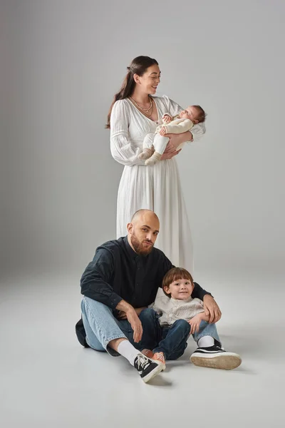 Padre barbudo sentado en el suelo con su pequeño hijo alegre al lado de su esposa con su bebé - foto de stock