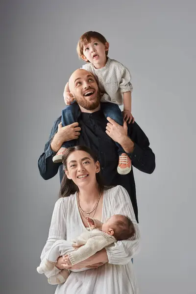 Alegre madre sentada con el bebé con su marido sosteniendo a su pequeño hijo en hombros detrás de ella - foto de stock
