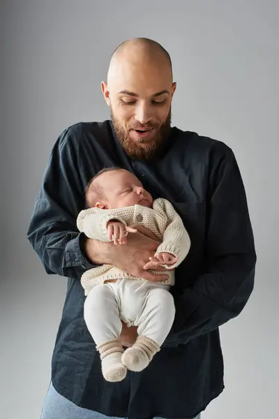 Joyous father in stylish attire holding his sleeping newborn baby on gray backdrop, family concept — Stock Photo