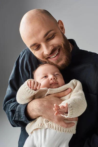 Tiro vertical de padre barbudo alegre abrazando a su hijo recién nacido en un cálido cárdigan, concepto familiar - foto de stock