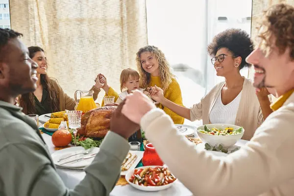 Alegres amigos multirraciales y familiares tomados de la mano y orando juntos en la mesa de Acción de Gracias - foto de stock