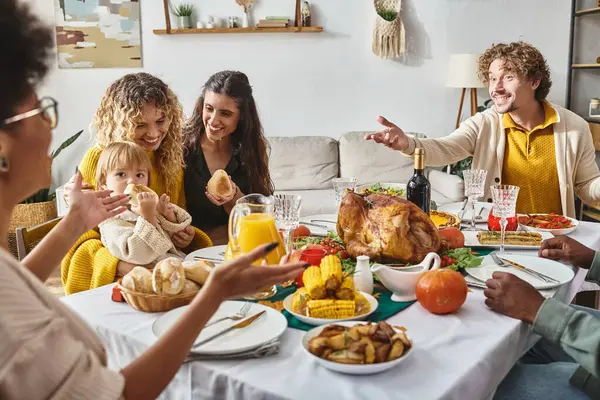 Feliz familia multirracial hablando y haciendo gestos durante la cena de Acción de Gracias al lado del pavo asado - foto de stock