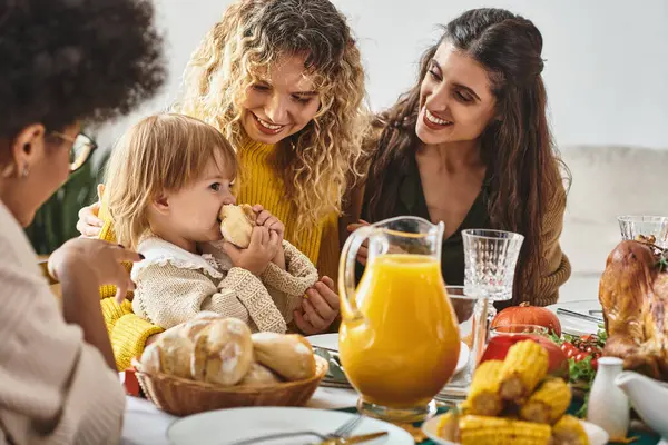 Happy multiethnic women looking at toddler girl eating bun during Thanksgiving celebration — Stock Photo