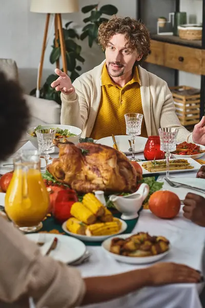 Hombre rizado haciendo gestos y hablando durante la cena de Acción de Gracias en círculo familiar, pavo asado en la mesa - foto de stock