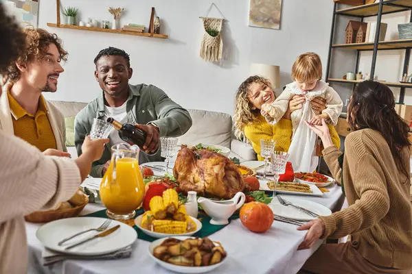Hombre afroamericano vertiendo vino en el vaso, familia multiétnica celebrando Acción de Gracias en casa - foto de stock